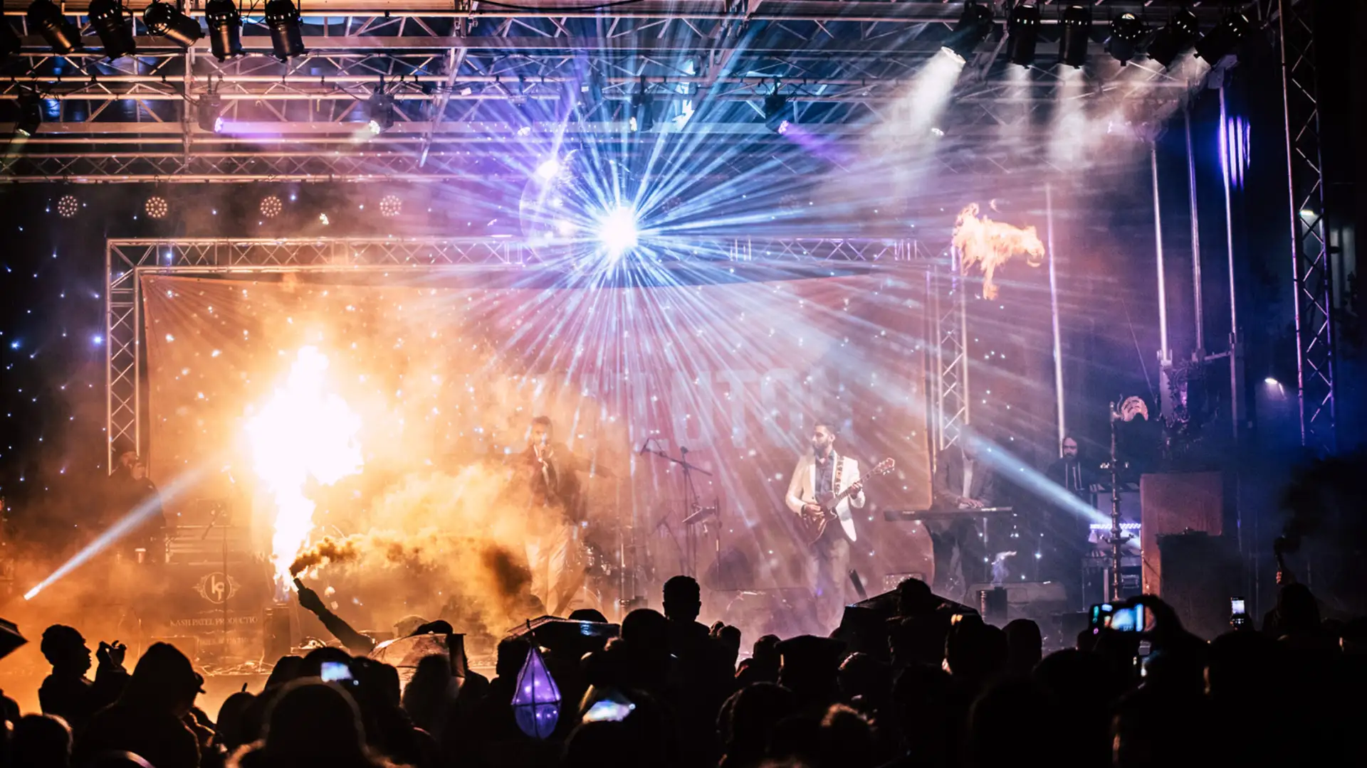 Band performing on a stage lit with coloured lights and sparks in front of a crowd